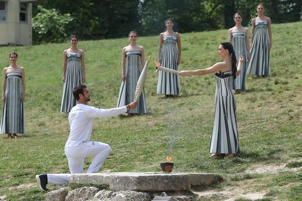 扮演最高女祭司的演員瑪麗·米娜（前右）點(diǎn)燃第一棒火炬手——希臘運(yùn)動(dòng)員、東京奧運(yùn)會(huì)男子賽艇項(xiàng)目金牌得主斯特凡諾斯·恩圖斯科斯手中的火炬。新華社記者 趙丁喆 攝