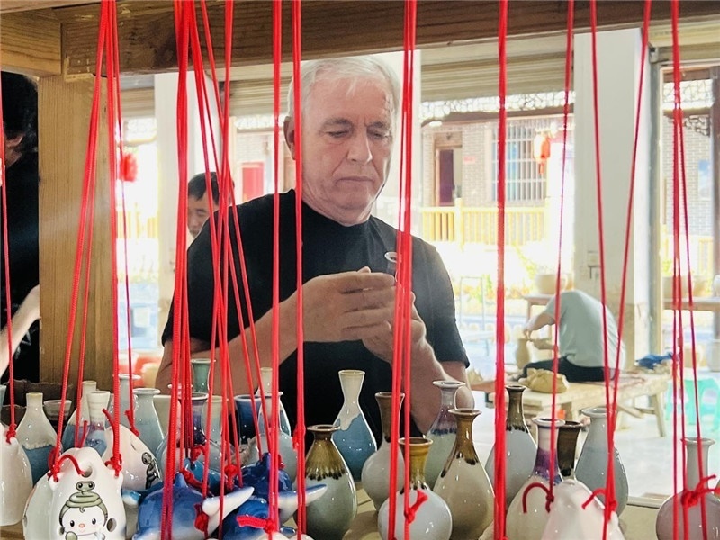 鄉(xiāng)愁如同一根根紅線，將游子與故鄉(xiāng)永遠(yuǎn)地牽連到一起?；逄账囄幕瘎?chuàng)意一條街的母氏陶藝館內(nèi)，紅線吊著陶瓶，思緒游走萬里。摩爾多瓦“信息標(biāo)簽”通訊社社長亞歷山大·塔納斯端詳著館內(nèi)展示的陶藝作品，并挑選出心儀的小擺件帶回摩爾多瓦，也帶走花茂村的“鄉(xiāng)愁”。（張亞欣攝）